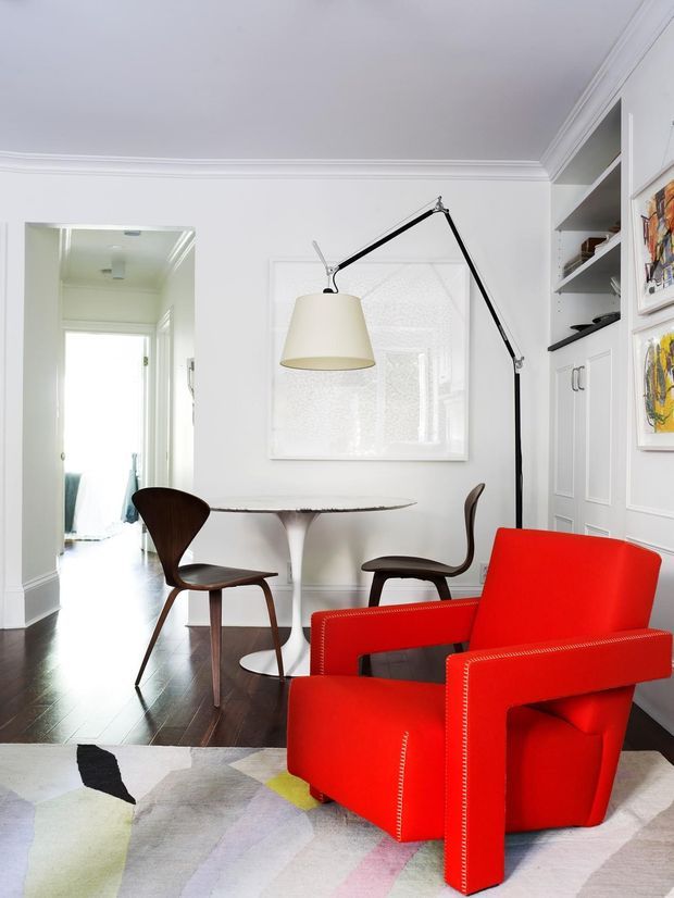 Bright, refined dining and seating area in a white-trimmed interior. A vivid red upholstered armchair sits in the foreground on a pale abstract rug, while a sculptural round dining table with dark wood chairs occupies the adjoining space. Built-in white shelving and panelled cabinetry line the right wall, with a large articulated floor lamp and artwork adding visual interest. Dark timber flooring, decorative ceiling mouldings, and a bright hallway contribute to the airy yet high-contrast composition.