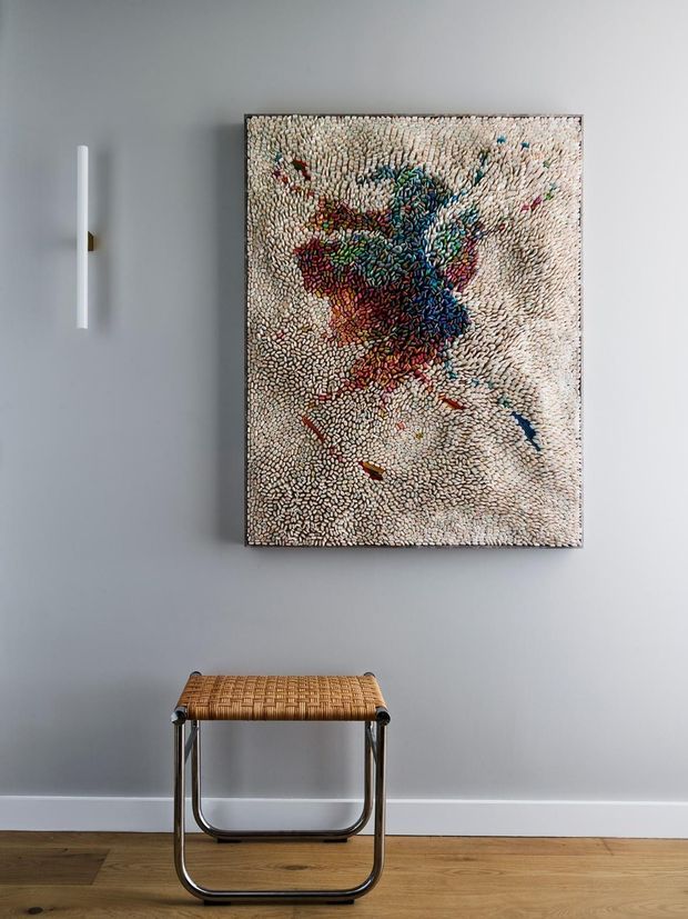 A minimalist interior vignette featuring a framed abstract artwork mounted on a pale grey wall, a slim white wall light, and a small woven-seat stool with a tubular metal frame. The space has warm timber flooring, white skirting boards, and a restrained, contemporary presentation.