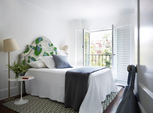 Bright, compact bedroom with white walls and ceiling trim, a white upholstered bed dressed in white bedding, and a bold green-and-white patterned headboard. A grey throw and cushion add contrast, while plantation shutters frame open balcony doors overlooking greenery. The room includes bedside tables, lamps, a patterned rug, and a dark wood console or dresser in the foreground.
