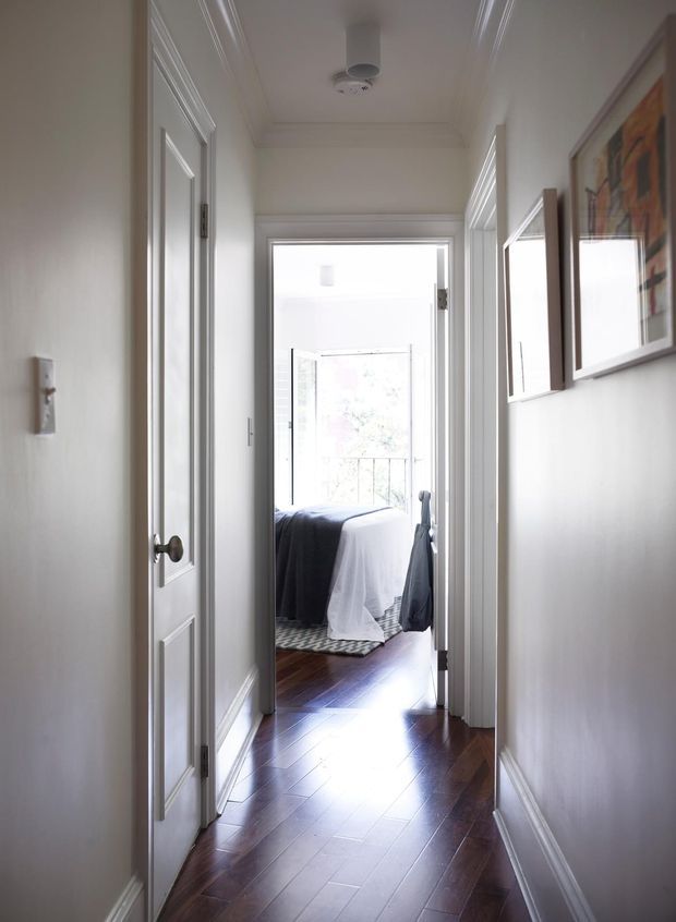 A bright, narrow residential hallway with dark polished timber flooring, white painted walls, decorative trim and panelled interior doors. Framed artwork hangs along the right wall, and the corridor leads to a sunlit bedroom with a partially visible bed and balcony or window beyond.