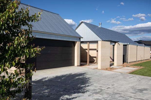 Exterior view of a contemporary single-storey residential property with a large attached garage, pale masonry walls, dark timber garage door, and steep corrugated metal gabled roofs. A broad concrete driveway and paved pedestrian path lead through a landscaped suburban setting.