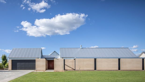 A wide, low-slung modern residential exterior viewed straight on, with a long corrugated metal pitched roof, pale horizontal timber-clad walls, dark vertical framing, a recessed timber entry door, and a large manicured lawn in front. The composition is clean and minimalist beneath a bright blue sky.