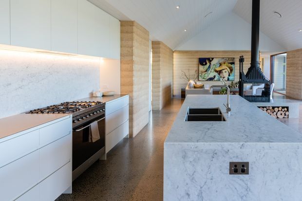Contemporary open-plan kitchen with a large white marble island incorporating a double sink and seating area, pale cabinetry, a gas cooktop, and a marble splashback. Timber-clad structural columns and walls, a pitched white-lined ceiling with recessed lighting, polished concrete flooring, and a black suspended fireplace contribute to the refined interior. A living area and artwork are visible beyond the kitchen.