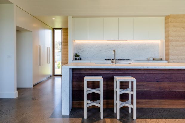 A bright contemporary kitchen with a long white island and dark timber front, two stools, white overhead cabinetry, a pale stone splashback, integrated under-cabinet lighting, and polished concrete flooring. A hallway and glazed exterior door are visible beyond the kitchen.