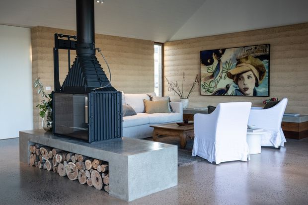 A contemporary open-plan living room with a prominent black wood-burning fireplace and exposed chimney, set into a concrete hearth with a decorative stacked-log niche. The room features textured tan walls, polished concrete flooring, a light sofa, slipcovered armchairs, a timber coffee table, and a colourful framed artwork.