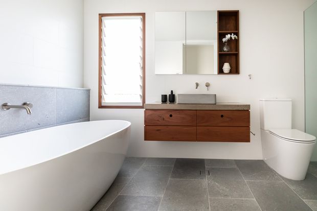 Bright contemporary bathroom featuring a freestanding white bathtub, wall-mounted timber vanity with a grey stone benchtop, integrated basin, mirrored wall cabinet, louvred timber-framed window, wall-mounted taps, white toilet, and large grey floor tiles. The palette is neutral with warm timber accents and abundant natural light.