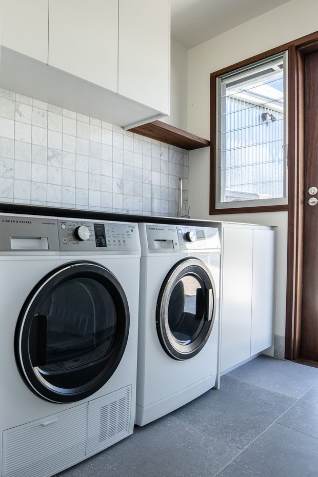 A bright, compact laundry room with front-loading white washer and dryer beneath white overhead cabinetry. The room features a pale square tiled splashback, dark grey tiled flooring, a timber shelf, timber-framed window, and dark timber door.