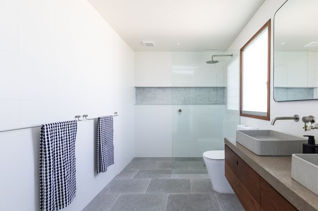 Bright contemporary bathroom with a walk-in glass shower, wall-mounted rain shower, freestanding toilet, twin vessel basins on a long timber vanity, large mirror, grey stone-look floor tiles, and a pale grey tiled shower wall. Timber-framed window and coordinated patterned towels add warmth and contrast.