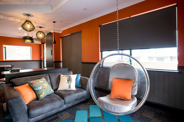 A vibrant contemporary living room with a dark grey sectional sofa, hanging transparent bubble chair, orange and teal cushions, dark wall panelling, and vivid burnt-orange walls. Large roller blinds cover the windows, while sculptural pendant lights and a layered ceiling add visual interest. A small workspace or desk area is visible at the rear.