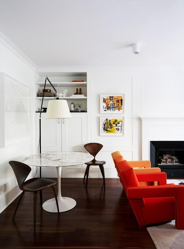 Bright, refined living and dining area with white paneled walls, built-in open shelving and cabinetry, dark timber flooring, a round marble-topped pedestal dining table, sculptural dark wood dining chairs, and vivid red-orange lounge chairs arranged around a black fireplace. Abstract artwork and a large floor lamp add visual interest.