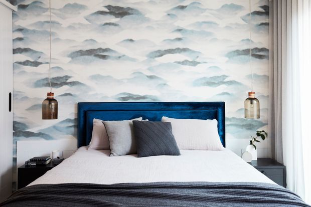 A stylish bedroom centered on a neatly made bed with a deep blue upholstered headboard, layered white and grey bedding, and charcoal throw. The wall behind the bed features a soft blue-grey cloud or abstract mountain mural, complemented by symmetrical hanging pendant lights, mirrored wardrobe panels, dark bedside tables, and sheer curtains.