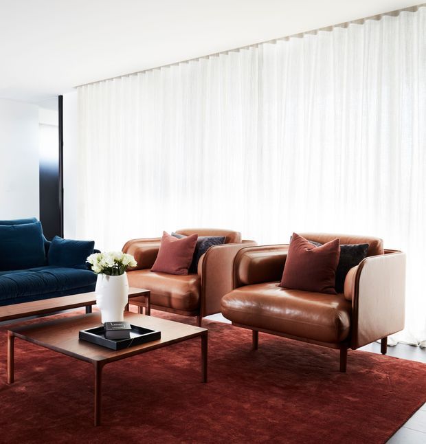 Bright, refined living room with floor-to-ceiling sheer curtains, a deep blue sofa, two cognac leather lounge chairs, a dark red area rug, and a low timber coffee table. The seating arrangement creates a warm, contemporary sitting space with strong contrast between the white backdrop, rich leather, blue upholstery, and burgundy rug.