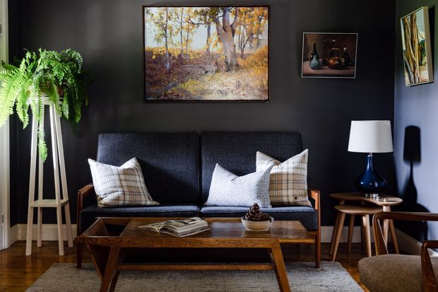 A dark, inviting living room featuring a mid-century-style charcoal sofa with plaid and textured cushions, a low timber coffee table, and a sculptural side table with a blue ceramic lamp. Black walls display framed artwork, while a large fern on a timber plant stand adds greenery. Timber flooring and a textured neutral rug complete the warm, layered interior.