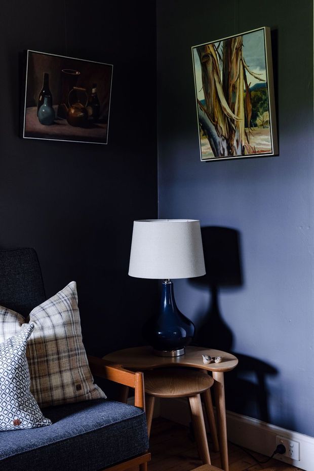 A moody, intimate corner of a living room featuring a dark blue upholstered armchair with patterned and plaid cushions, a mid-century-style wooden side table, and a dark ceramic lamp with a white fabric shade. Deep navy and charcoal walls are decorated with framed artwork, while dramatic directional lighting creates strong shadows.