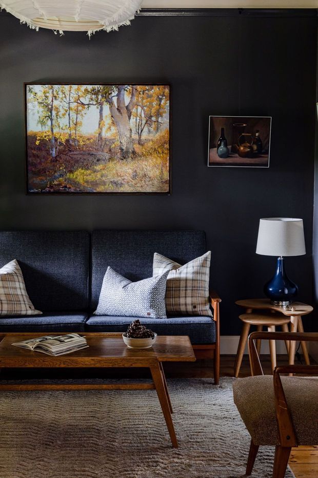 A warmly styled mid-century-inspired living room with a dark charcoal feature wall, deep navy sofa, timber coffee table, side tables, and an upholstered armchair. Patterned cushions, framed artwork, a blue ceramic lamp, books, and a textured neutral rug create a refined, cozy atmosphere. Timber flooring and a large woven pendant light are visible.