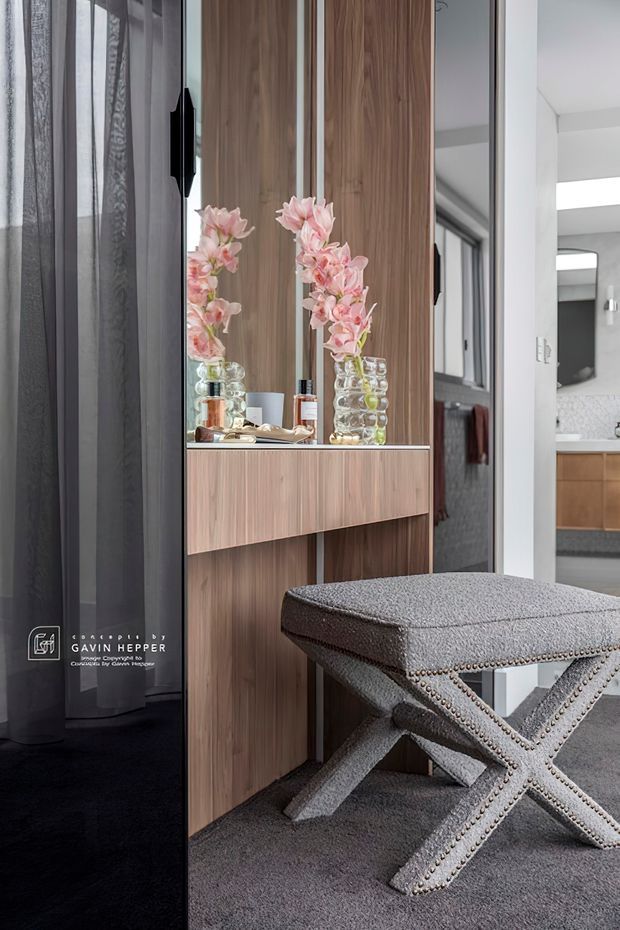 A refined interior vignette featuring a tall walnut-toned built-in dressing or entry console with mirrored panels and a floating drawer. Pink floral arrangements and decorative objects sit on the console, while a grey upholstered X-frame bench occupies the foreground. Dark sheer curtains and carpet contrast with the light modern hallway and bathroom visible beyond.