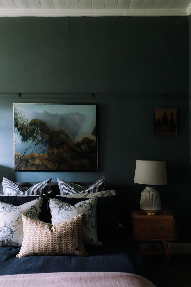 A moody, richly layered bedroom with a dark green wall, white-painted paneled ceiling and trim, and a bed dressed in multiple patterned cushions and dark blue bedding. A landscape artwork hangs above the bed, complemented by a smaller framed artwork and a sculptural table lamp on a mid-century-style wooden bedside table.