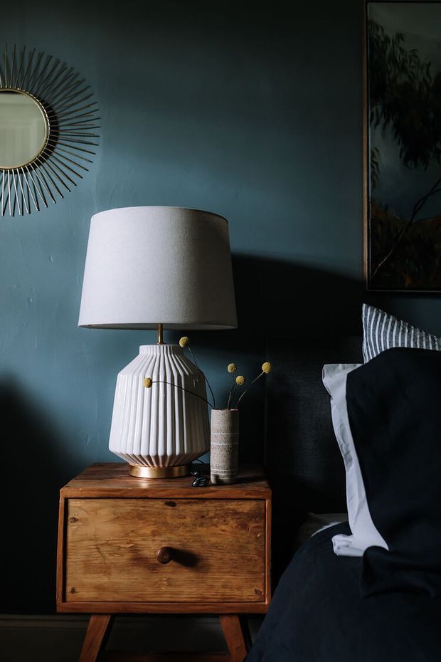 Moody close-up of a bedroom bedside setting with a dark teal painted wall, black upholstered bed, white and black bedding, and a warm timber bedside table. A sculptural pleated ceramic lamp, framed artwork, sunburst mirror, and small decorative vase create a refined, eclectic atmosphere with strong side lighting and deep shadows.