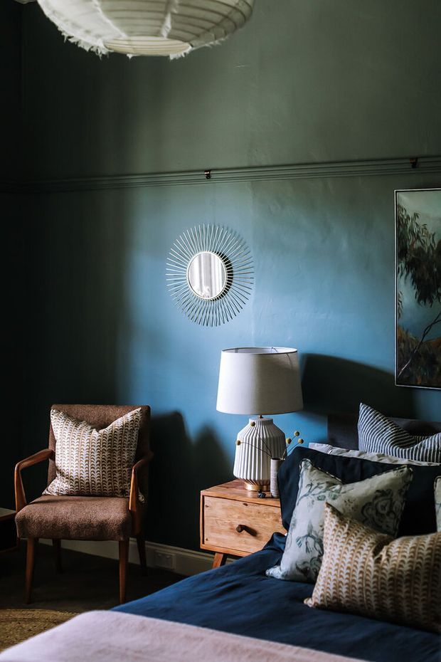 Moody, richly layered bedroom interior with deep blue-green walls, a mid-century timber bedside table, upholstered wooden armchair, patterned cushions, a blue bedspread, framed artwork, and a sunburst wall mirror. A woven pendant light and warm timber accents add texture and character.