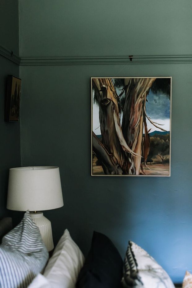 A moody bedroom corner with deep blue-green painted walls, decorative wall moulding, framed tree artwork, and a bedside table topped with a white ceramic lamp. Layered pillows and bedding occupy the foreground, creating a dark, intimate, eclectic atmosphere.