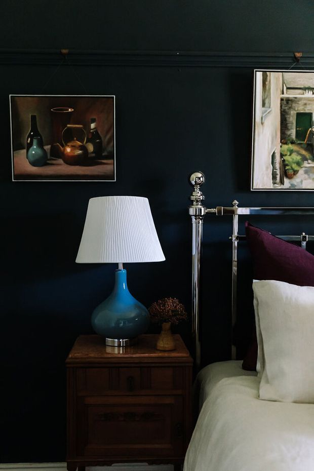 A moody, richly styled bedroom vignette featuring a dark navy or charcoal wall, a traditional dark timber bedside table, a blue ceramic table lamp with a pleated white shade, and part of a polished metal bed frame with white bedding and a deep burgundy cushion. Framed still-life and landscape artwork add a classic, curated character.