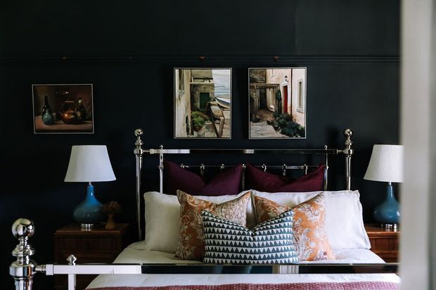 A richly styled bedroom with a black accent wall, polished metal four-poster bed, layered white bedding, and multiple patterned burgundy, teal, and orange cushions. Matching bedside tables hold blue glass lamps with white shades, while framed artwork decorates the wall.