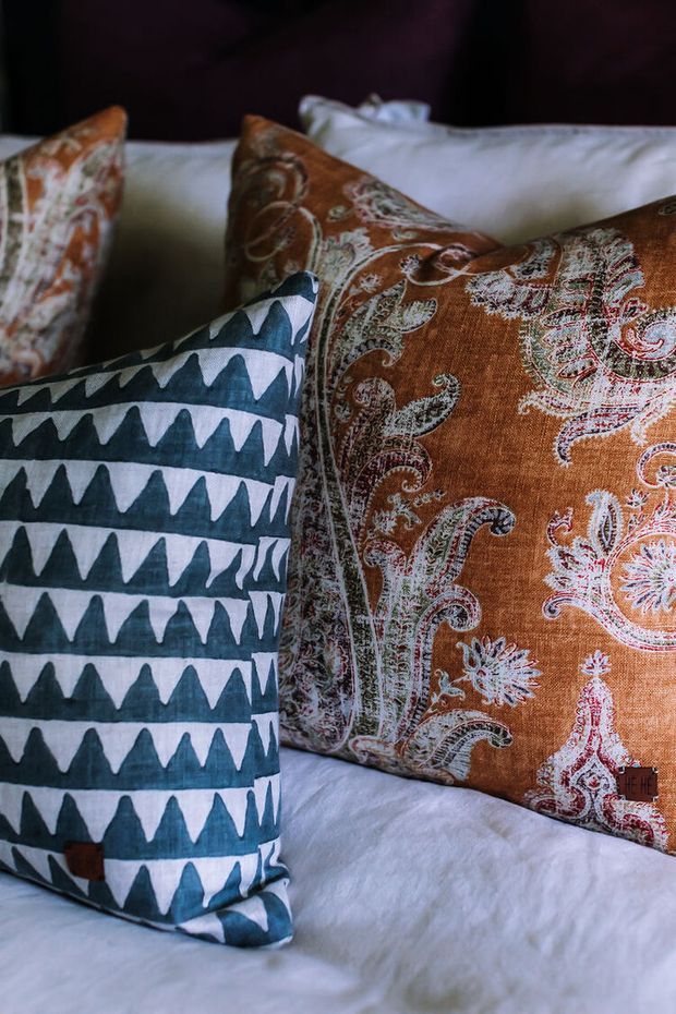 Close-up of layered decorative cushions on a bed or upholstered seating, featuring a bold geometric blue-and-white pattern in the foreground and richly patterned rust-orange paisley cushions behind. The image is a detailed textile and soft-furnishing vignette with a warm, eclectic character.