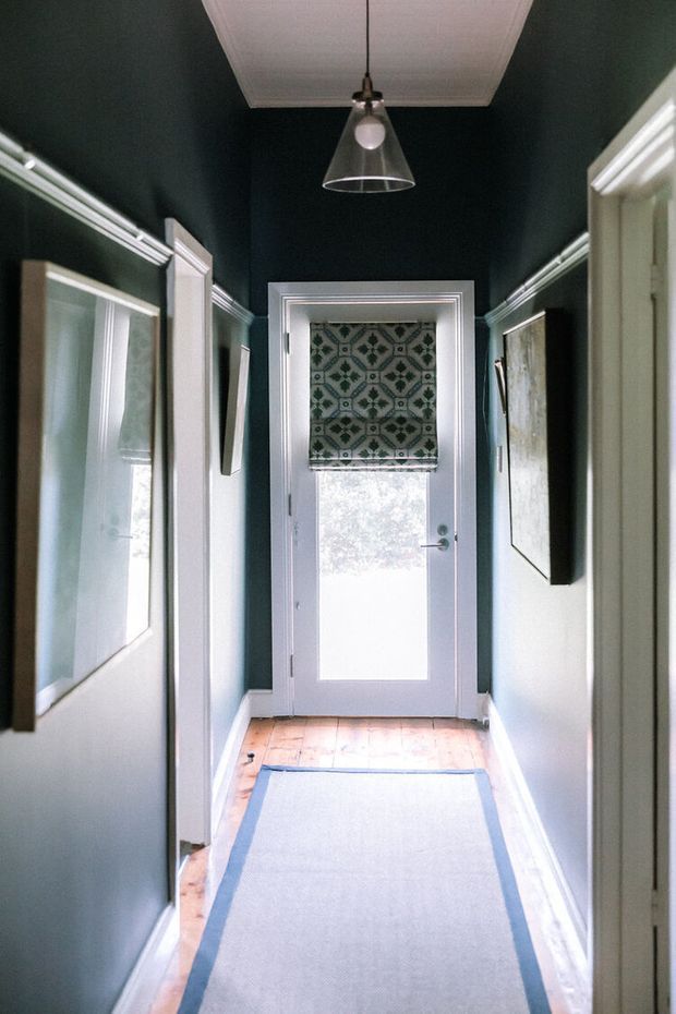 A narrow, richly coloured hallway with deep green walls, white decorative trim and door frames, a light timber floor, and framed artwork. A patterned roller blind covers the upper portion of a glazed exterior door at the end of the corridor. A simple pendant light hangs from a white coffered ceiling, while a long pale runner with a blue border extends through the space.