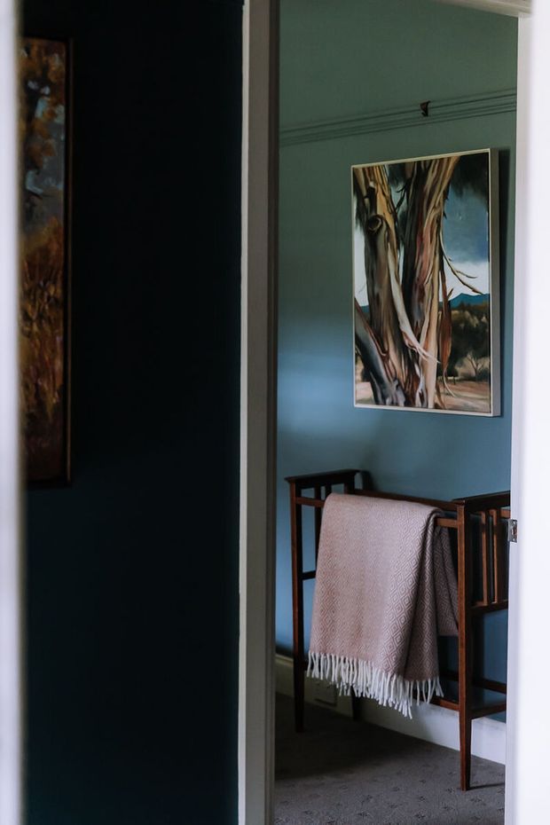 A view through a doorway into a compact, traditionally detailed interior space with deep blue-green painted walls, decorative picture-rail moulding, dark timber furniture, and a patterned fringed throw draped over a wooden bench or chair. A framed tree landscape artwork provides a natural focal point, while dark carpet and white trim complete the moody, refined setting.