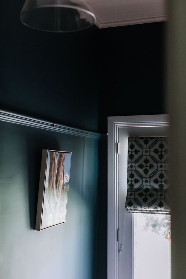 A moody interior detail showing a deep teal painted wall with decorative white dado-style moulding, a framed artwork, a ceiling light, and a white-trimmed glazed door or window fitted with a patterned Roman blind.