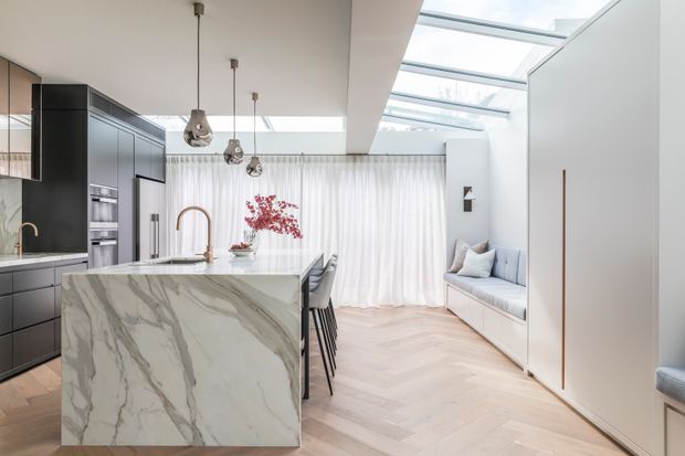 Bright contemporary open-plan kitchen with a large veined stone island, dark built-in cabinetry, integrated appliances and pendant lights. A skylit seating nook with built-in bench cushions sits beyond the kitchen, with sheer curtains and pale herringbone timber flooring creating an airy, refined interior.