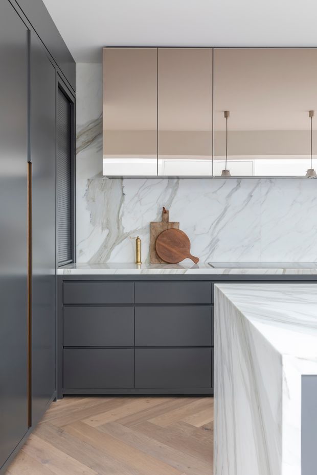 A refined contemporary kitchen with dark charcoal full-height cabinetry, pale wood-toned upper cupboards, and dramatic white veined stone used across the splashback and island. Brass tapware and slim pendant lights add warm accents, while the timber herringbone floor softens the monochrome palette.