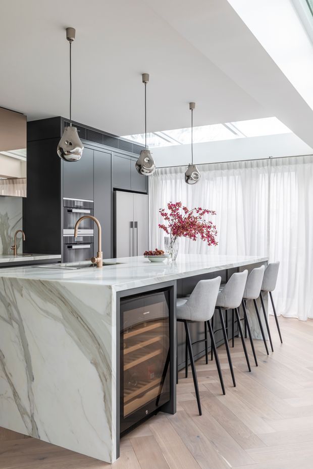 Bright contemporary kitchen with a large veined white stone waterfall island, dark grey full-height cabinetry, integrated appliances, wine fridge, and a bronze-toned faucet. Four upholstered bar stools line the island beneath reflective pendant lights. A roof skylight and full-height sheer curtains provide abundant natural light, with pale timber flooring throughout.