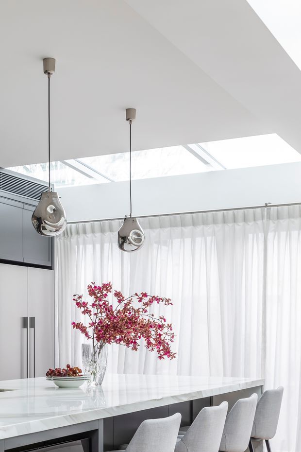 Bright contemporary kitchen and dining area with a large white stone island, integrated pale cabinetry, grey upholstered bar stools, sculptural chrome pendant lights, sheer curtains, and a glazed skylight or clerestory above. A floral arrangement and fruit bowl sit on the island.