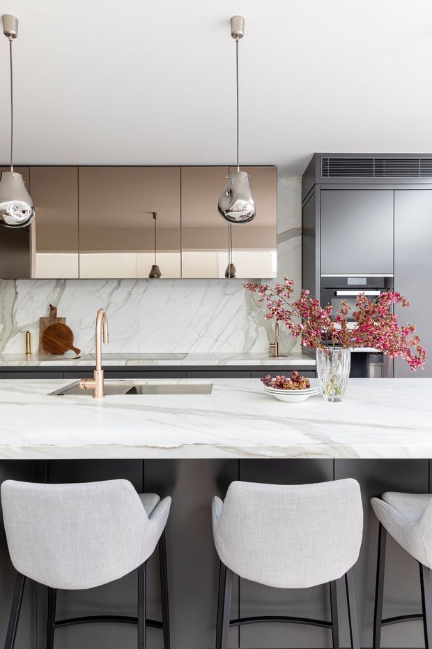 Contemporary kitchen featuring a large white marble island with integrated sink and rose-gold tapware, dark cabinetry, mirrored bronze-toned overhead cabinetry, full-height marble splashback, integrated appliances, and sculptural pendant lights. Light upholstered bar stools line the island, with floral branches adding a soft decorative accent.