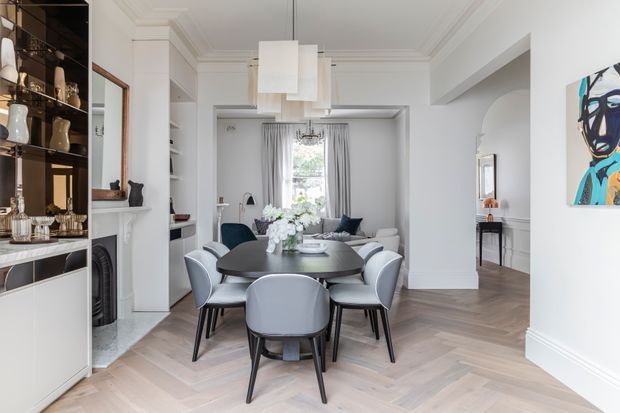 A refined open-plan dining area with a large round dark dining table surrounded by upholstered grey chairs, positioned between built-in display cabinetry and a bright living space. The room features white walls, ornate ceiling mouldings, herringbone timber flooring, a sculptural pendant light, a fireplace surround, and contemporary artwork.
