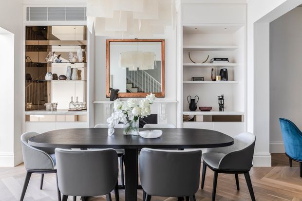 A refined dining room with a large dark oval dining table surrounded by upholstered grey chairs. Built-in white shelving and display cabinetry line the rear wall, with decorative objects and glassware. A timber-framed mirror reflects a staircase, while a sculptural pendant hangs above the table. The room has light walls, herringbone timber flooring, and a bright, elegant contemporary-classic character.