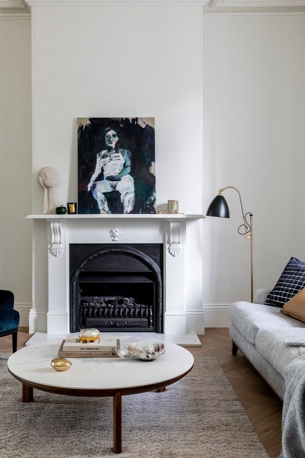 A refined living room centered on a classic white mantel fireplace with decorative corbels and a dark arched firebox. The room features pale panelled walls, a large abstract artwork above the mantel, a sculptural floor lamp, a light grey sofa with patterned and tan cushions, a deep teal armchair, and a round white coffee table over a textured neutral rug. The overall look combines traditional architectural detailing with contemporary furnishings.