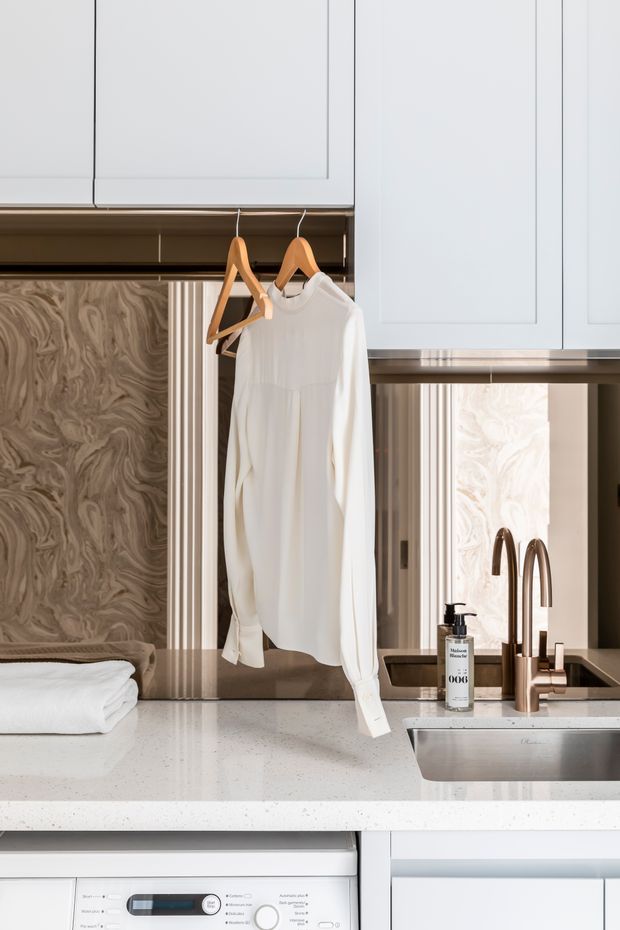 Close-up of a refined laundry room with pale cabinetry, a white stone-look benchtop, stainless-steel sink, warm metallic gooseneck tapware, integrated washing machine, and hanging laundry. A vertically fluted and marbled feature backsplash adds texture behind the work area.