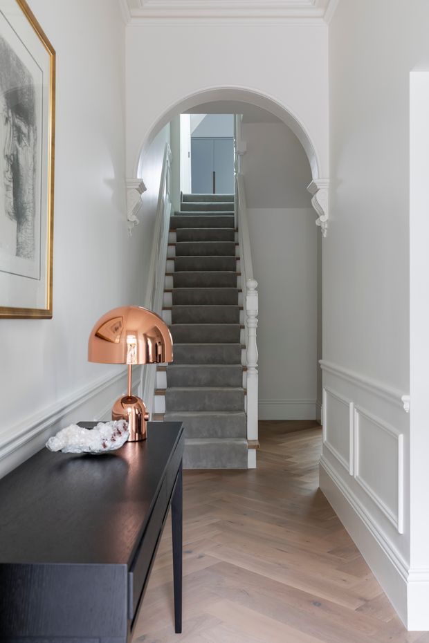 A refined residential hallway with a central staircase leading through a decorative arched opening. The space features white walls with traditional panel moulding, ornate arch detailing and cornices, a grey carpeted stair, pale timber herringbone flooring, and a dark console with copper-toned lamps. A framed artwork adds visual interest to the left wall.