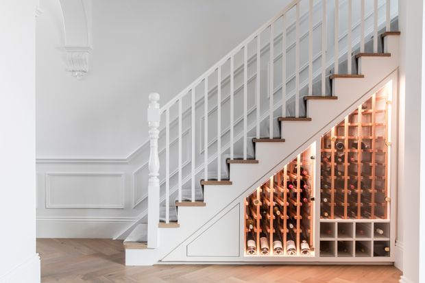 Bright, refined hallway or entry area featuring a white-painted staircase with slim vertical balustrades, dark timber stair treads, decorative wall panelling, and a built-in wine storage display beneath the stairs. The floor is laid in a light herringbone timber pattern, and warm lighting highlights the wine collection.