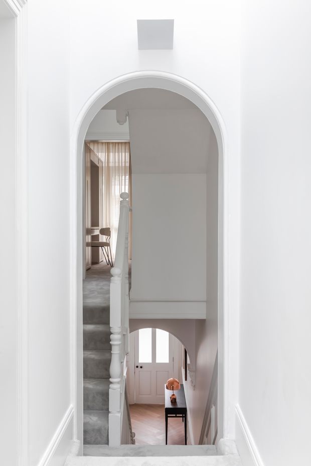 A bright, refined view through a white arched opening into a multi-level stair hall. The staircase has grey carpeted steps, a painted white turned balustrade, decorative wall mouldings, and a lower-level entry with pale pink double doors, timber flooring, and a small console table with a lamp. A glimpse of a warm-toned sitting or study area is visible at the upper landing.