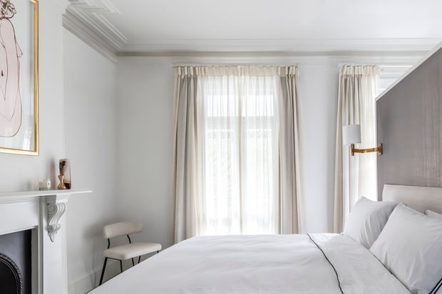 A bright, refined bedroom with a large upholstered bed dressed in white bedding, tall sheer-curtained windows, white walls, ornate ceiling cornices, and a decorative white fireplace mantel. A brass wall light and minimalist artwork add subtle accents, creating an elegant modern-classic interior.