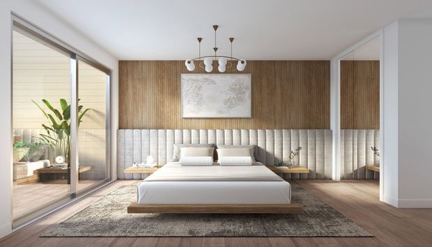 A bright, spacious contemporary bedroom with a low-profile upholstered bed centered against a full-width vertical timber feature wall. The wall includes a tall padded headboard, framed artwork, and integrated bedside surfaces. Warm timber flooring, a large neutral rug, soft layered bedding, sculptural pendant lighting, and full-height sliding glazing create an airy, refined retreat. A planted outdoor area is visible through the glazing and reflected in the adjacent mirror.
