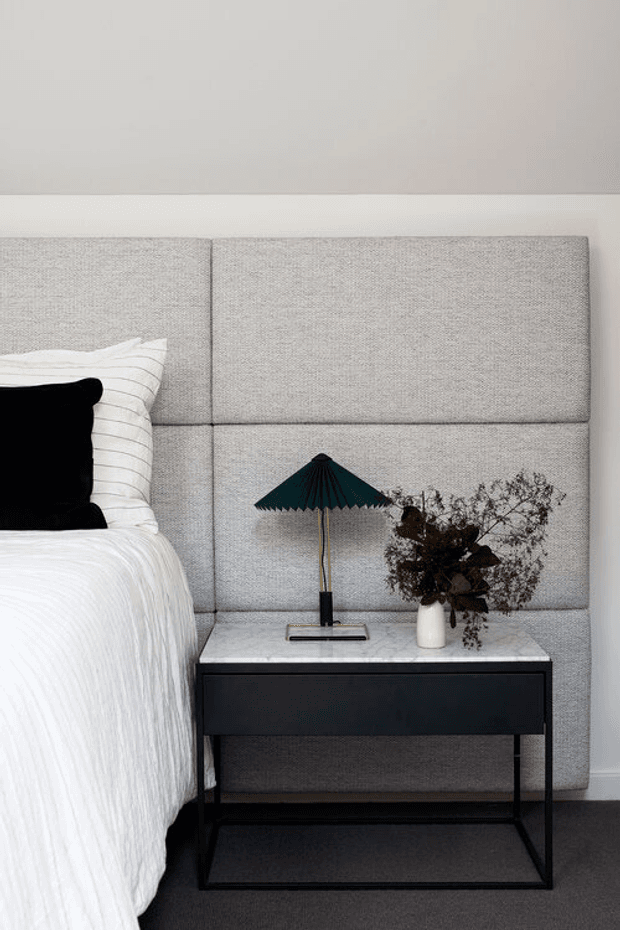 A close-up of a refined bedroom corner featuring a large light-grey upholstered, panelled headboard, white bedding, a black bedside table with a pale stone top, a dark pleated table lamp, and a white vase with dried foliage. The palette is restrained and monochromatic with soft textures and dark accents.