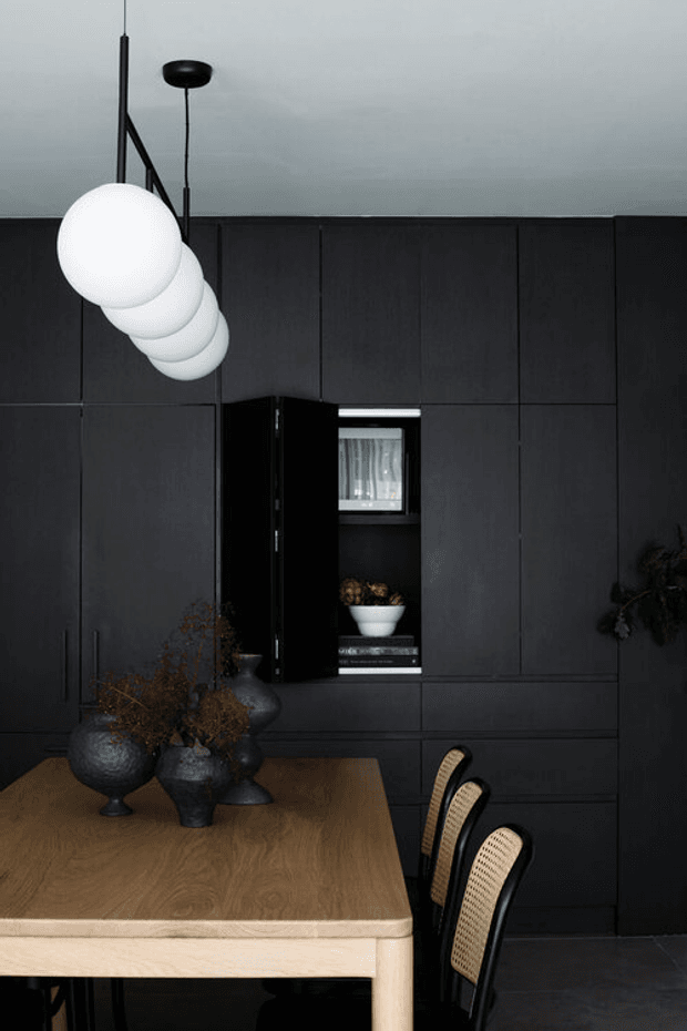 A dark, refined dining area with a large natural timber dining table and black cane-back chairs in the foreground. The back wall is fully fitted with matte charcoal-black cabinetry and integrated open shelving, while a sculptural cluster of white globe pendant lights hangs overhead. Black ceramic vases and dried branches provide restrained decoration, with a small amount of greenery at the side.