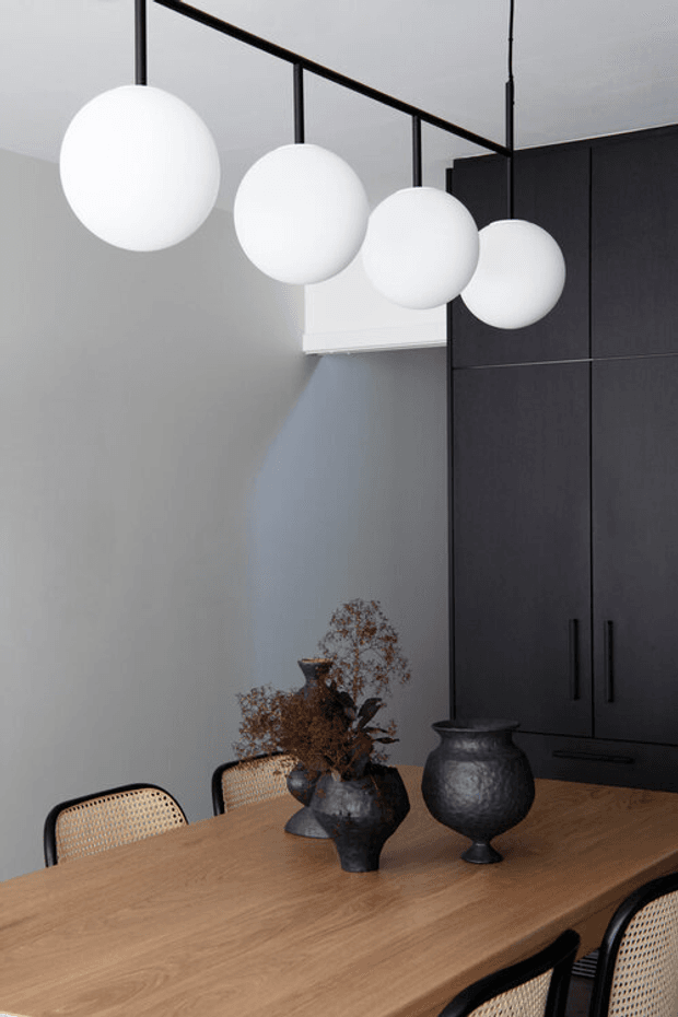 A refined dining area with a large light-wood dining table, black-framed cane chairs, sculptural dark decorative vessels, matte dark built-in cabinetry, and four spherical white pendant lights suspended from a linear black ceiling frame. The room has pale grey walls and a restrained, contemporary monochromatic palette.