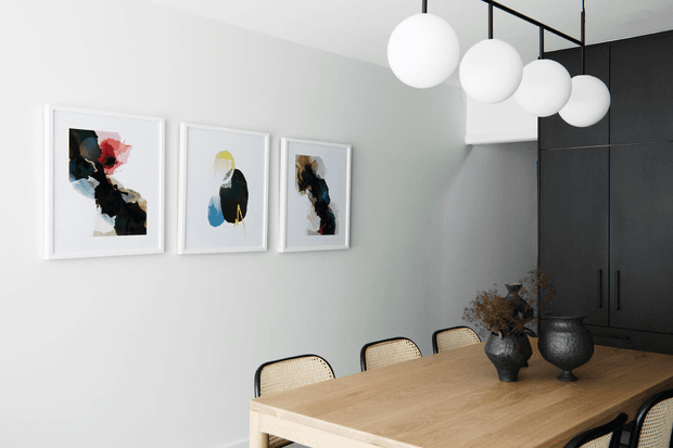 A contemporary dining area with a light timber dining table and woven cane dining chairs. The room has pale grey walls, dark built-in cabinetry, three abstract framed artworks, and a linear black ceiling light with multiple spherical white pendants. Dark sculptural vases with dried branches provide tabletop accents.