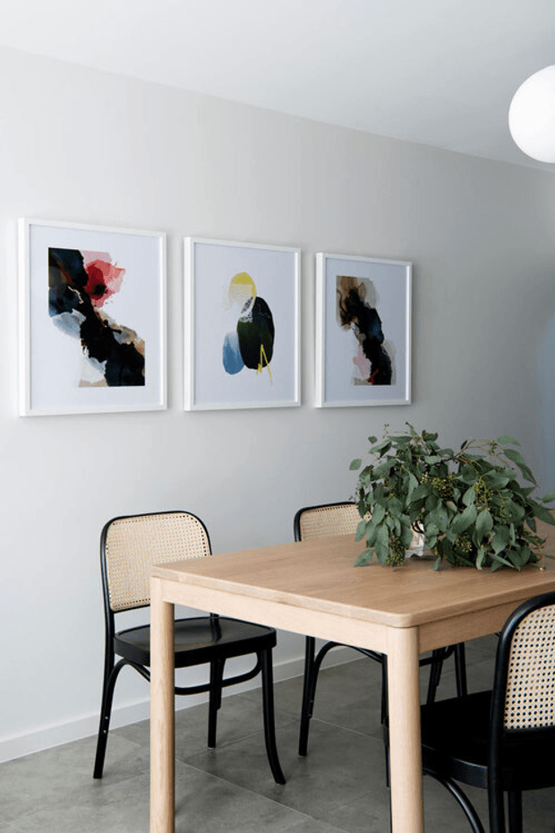 A bright, contemporary dining area with a light timber rectangular dining table, black-framed woven cane chairs, pale grey walls, grey tiled flooring, and three abstract artworks in white frames. A leafy plant forms a natural centrepiece, while a spherical white ceiling light is partially visible.