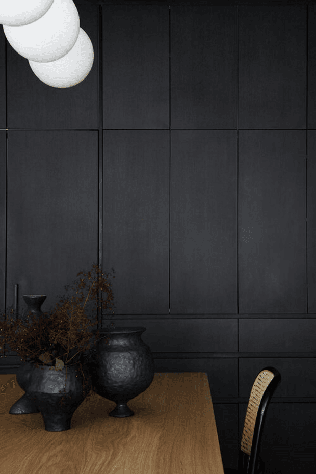 Moody, close-up interior detail of a dark panelled wall behind a warm timber dining table. Black sculptural ceramic vessels with dried branches sit on the table, accompanied by a black woven dining chair and cropped white globe pendant lights. The composition is refined and minimalist with strong contrast between the charcoal wall, timber surface and pale lighting.